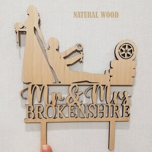 May include: A wooden wedding cake topper featuring a bride and groom with a car wheel and gear design. The words "Mr & Mrs BROKENSHIRE" are carved into the wood. The text "NATURAL WOOD" is above the figures.