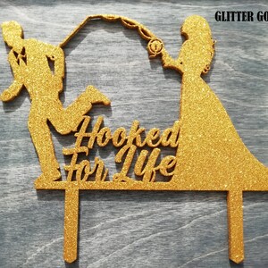 May include: A glitter gold cake topper featuring a silhouette of a couple, with the man running and the woman holding a fishing rod. The text "Hooked For Life" is in the center. The words "Glitter Gold" are in the top right corner.