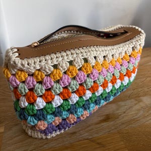 Può includere: Una borsa all'uncinetto multicolore con una cerniera in pelle marrone. La borsa presenta un arcobaleno di colori, tra cui viola, blu, verde, bianco, arancione, rosa e giallo. La borsa è fatta di filato e ha una chiusura con cerniera.