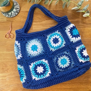 May include: A blue crocheted tote bag with a white and blue geometric pattern. The bag has two straps and is made of thick yarn.