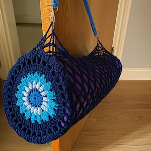 May include: A blue and white crocheted yoga mat bag with a purple yoga mat inside. The bag has a blue strap and a silver clip.
