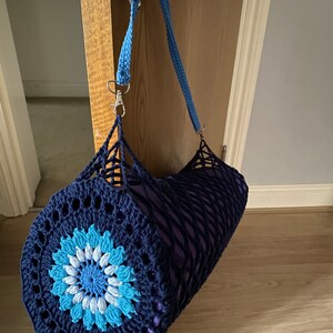 May include: A blue and white crocheted yoga mat bag with a flower design. The bag has a long blue strap and a silver clasp.