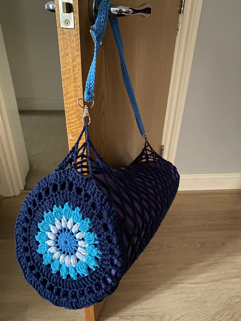May include: A blue and white crocheted yoga mat bag with a flower design. The bag has a long blue strap and a silver clasp.
