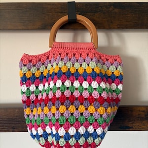 May include: A colorful, handmade crochet bag with a wooden handle. The bag features a vibrant pattern of red, pink, blue, green, yellow, and white stitches. The top edge is a solid pink color. The bag is hanging on a dark brown wooden bar.