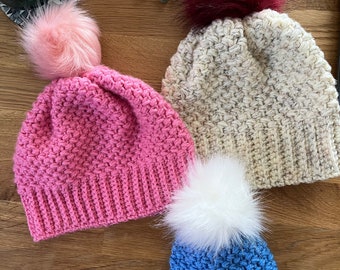 May include: Three crocheted hats with faux fur pom poms. The hats are in pink, beige, and blue. The pom poms are in pink, burgundy, and white.