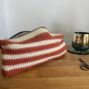 May include: A striped crocheted zippered pouch with a cream and rust color scheme. The pouch is lying on a wooden surface.