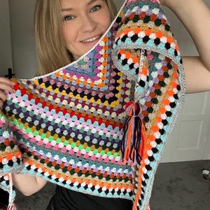 May include: A vibrant, hand-crocheted shawl in a granny square pattern. The shawl displays a spectrum of colours: orange, blue, green, and purple. It has a grey border and tassels. A unique, cosy accessory, perfect for adding a pop of colour.