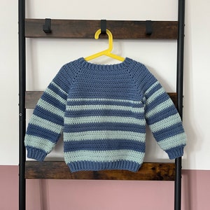 Könnte beinhalten: Ein gestrickter Pullover in Blau und Hellblau mit Streifen, der an einem gelben Kleiderbügel hängt.