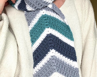 May include: A handmade crochet scarf with a chevron pattern. The scarf features stripes of white, teal, navy blue, and gray. The scarf is made of soft yarn and is designed to be worn around the neck.