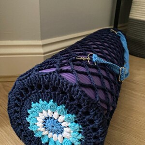 May include: A navy blue crocheted yoga mat bag with a blue and white flower design. The bag has a purple mesh lining and a blue strap with a silver clip.