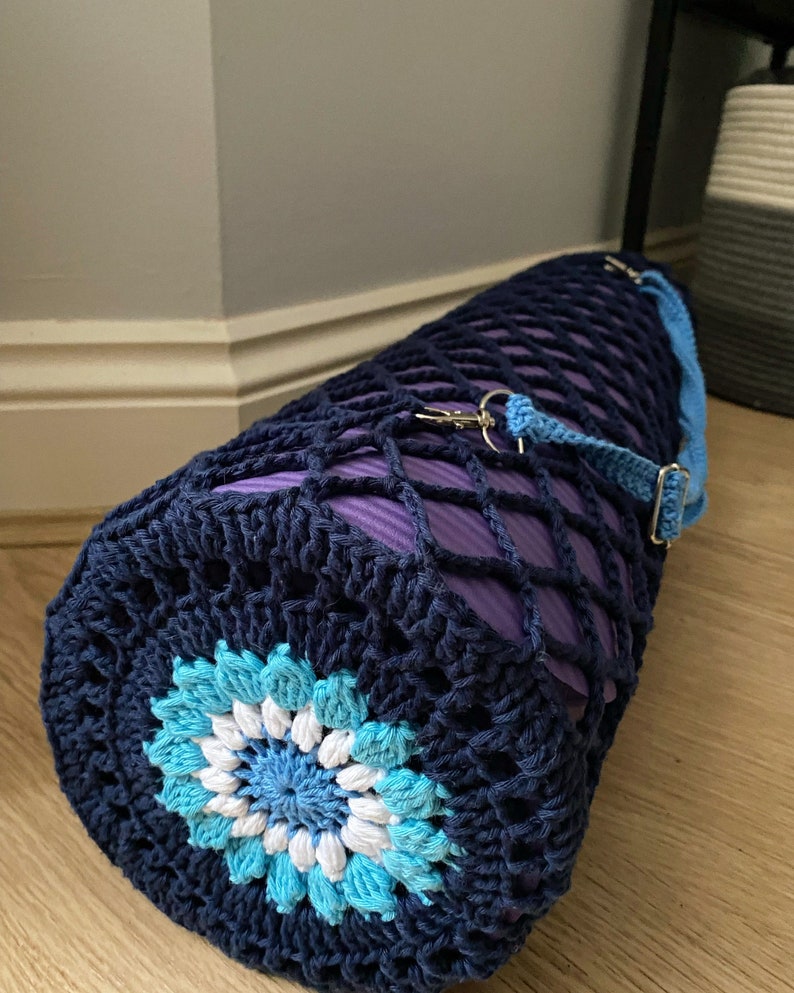 May include: A navy blue crocheted yoga mat bag with a blue and white flower design. The bag has a purple mesh lining and a blue strap with a silver clip.