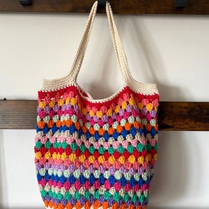 May include: A colorful, handmade crochet tote bag. The bag features a vibrant patchwork design with a variety of colors, including red, yellow, blue, green, and purple. The top and handles are a cream color. The bag is hanging on a wooden rack.