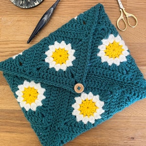May include: A teal crocheted pouch with a daisy pattern and a wooden button closure. The pouch is made with white and yellow yarn.