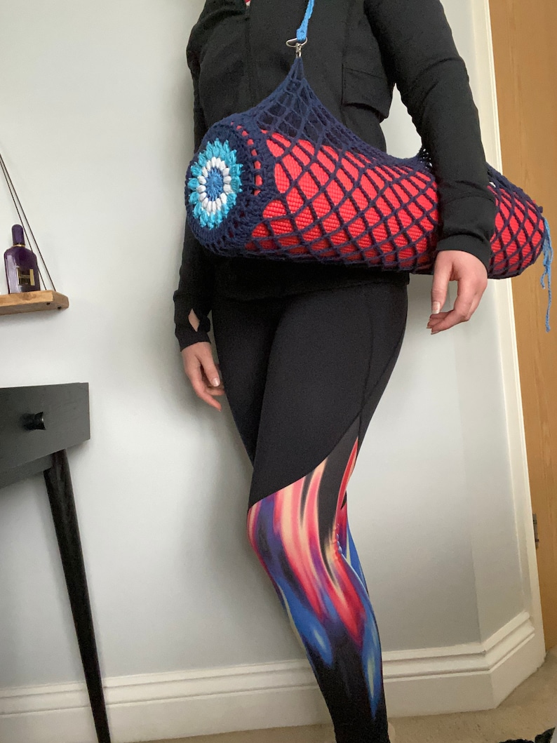 May include: A blue crocheted yoga mat bag with a colorful flower design. The bag is worn over the shoulder and holds a red yoga mat.
