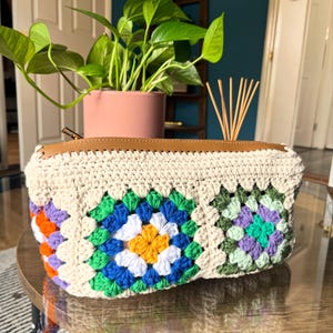 May include: A cream-colored crochet pouch with a brown leather top and zipper. The pouch features colorful granny square designs in shades of blue, green, orange, purple, and yellow. The pouch is sitting on a round, glass table.