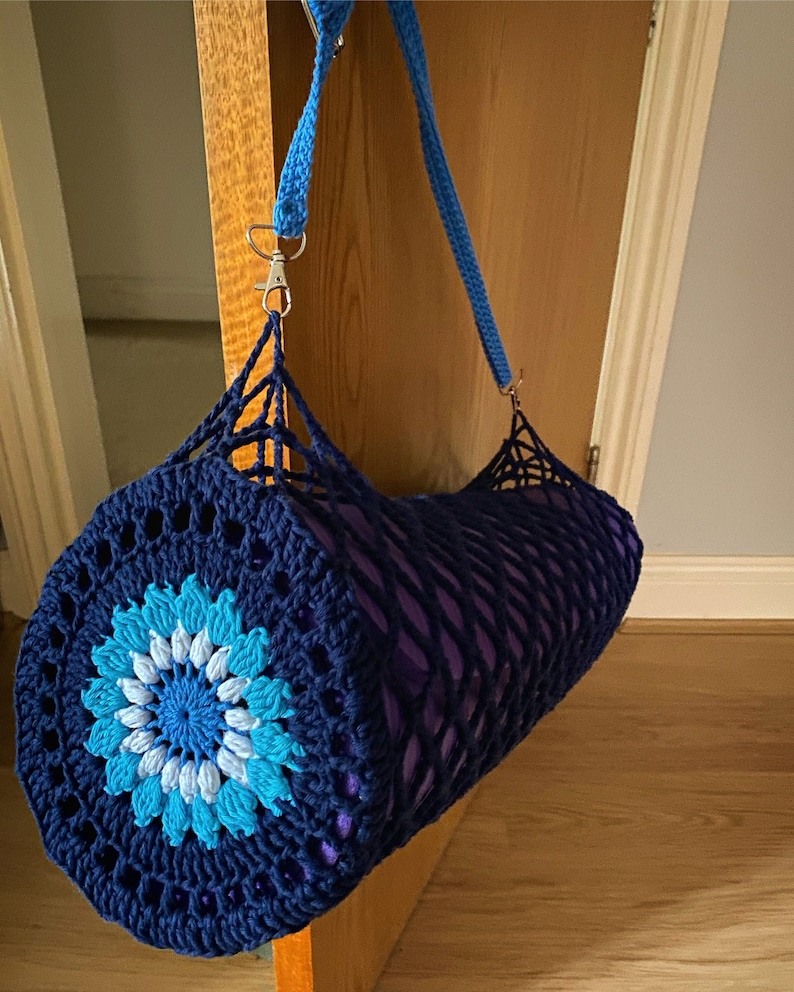 May include: A blue and white crocheted yoga mat bag with a purple yoga mat inside. The bag has a blue strap and a silver clip.