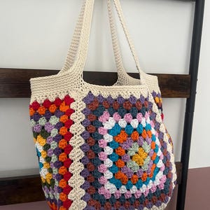 May include: A crocheted tote bag with a colorful granny square pattern in shades of red, orange, yellow, green, blue, purple, and pink. The bag has long, cream-colored straps.