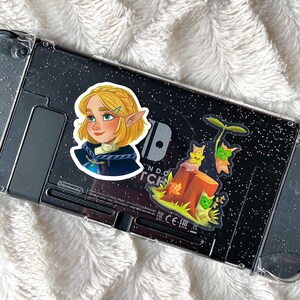 May include: Two stickers on a black and silver Nintendo Switch console. One sticker features a cartoon girl with blonde hair and a blue dress. The other sticker features a cartoon tree with a brown trunk and green leaves.