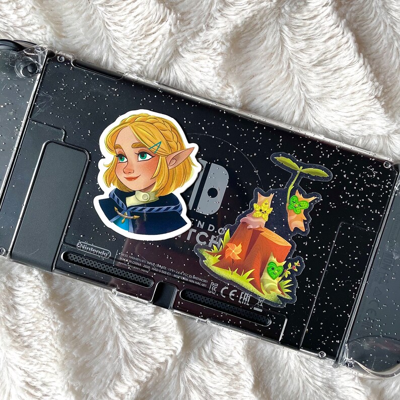 May include: Two stickers on a black and silver Nintendo Switch console. One sticker features a cartoon girl with blonde hair and a blue dress. The other sticker features a cartoon tree with a brown trunk and green leaves.