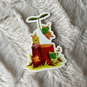 May include: A sticker featuring three cartoon animals with green leaves on their faces. The animals are sitting on a tree stump with a brown base and a green top. The background is a white surface.