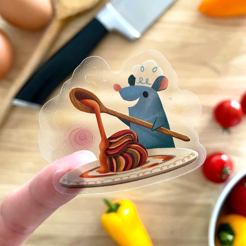 May include: A blue mouse wearing a chef's hat pours sauce from a wooden spoon onto a plate of colorful pasta. The mouse is smiling and holding the spoon. The plate has a gold rim and is decorated with a swirl pattern.