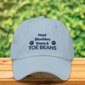 May include: A light blue baseball cap with the text "Head Shoulders Knees & TOE BEANS" embroidered in navy blue. Two paw prints are embroidered on either side of the text.