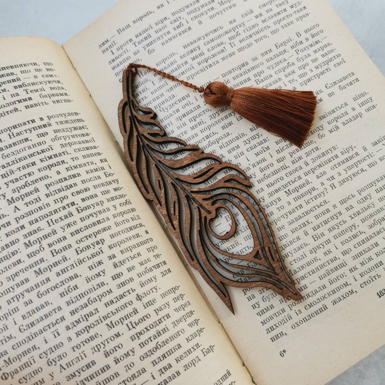 May include: A wooden bookmark shaped like a peacock feather with intricate cutouts. The bookmark has a brown tassel attached to the top.