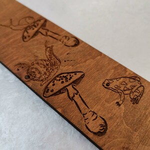 A wooden bookmark with a laser-engraved design of a frog, two snails, and mushrooms.