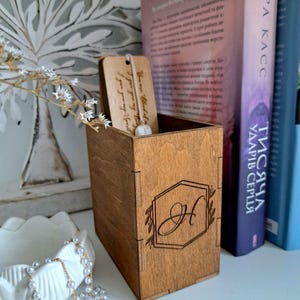 May include: A wooden box with an engraved letter "H" inside a hexagon, and a small wooden tag with the text "You are loved..." inside. A pearl necklace and books are in the background.