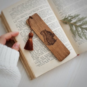 Könnte beinhalten: Holz-Lesezeichen mit detailliertem, graviertem Eichhörnchen-Design. Das Lesezeichen hat eine Quaste und liegt auf den Seiten eines offenen Buches. Das Holz hat einen warmen, natürlichen Farbton.