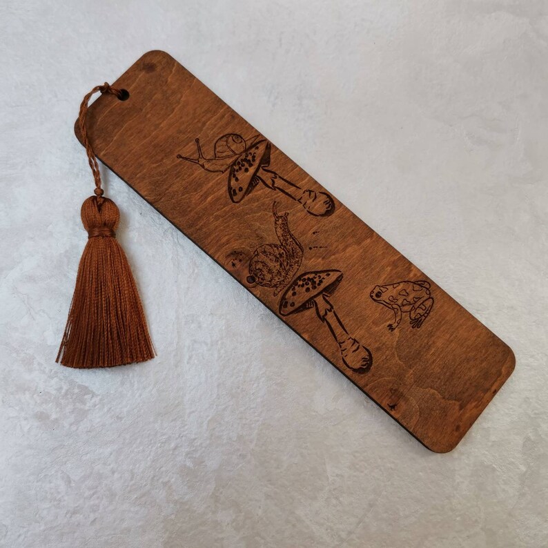 A wooden bookmark with a brown tassel. The bookmark is decorated with a laser-engraved design of a snail, a frog, and mushrooms.