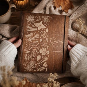 Peut inclure: Un livre de recettes carré en bois avec une finition brun foncé, présentant des illustrations gravées au laser de diverses herbes et épices sur le côté gauche et le texte "Galina recipe book" sur le côté droit, maintenu par un élastique.