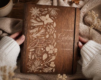 May include: A square wooden recipe book with a dark brown finish, featuring laser-engraved illustrations of various herbs and spices on the left side and the text "Galina recipe book" on the right side, secured with a vertical elastic band.