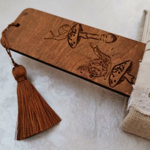 May include: A wooden bookmark with a brown tassel. The bookmark is engraved with two snails and mushrooms.