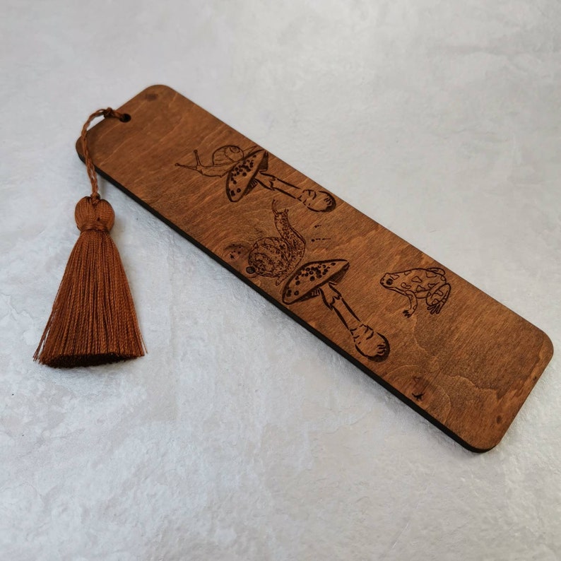 May include: A wooden bookmark with a brown tassel. The bookmark features a laser-engraved design of mushrooms, snails, and a frog.