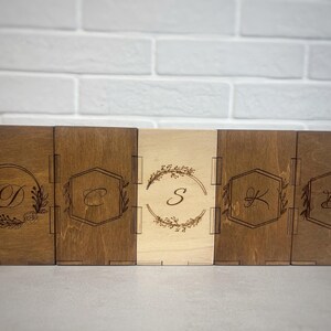 May include: Five rectangular wooden boxes arranged in a row, each featuring a laser-engraved monogram. The boxes vary in wood tone from light to dark brown. Each box displays a unique floral or geometric frame surrounding a single letter.