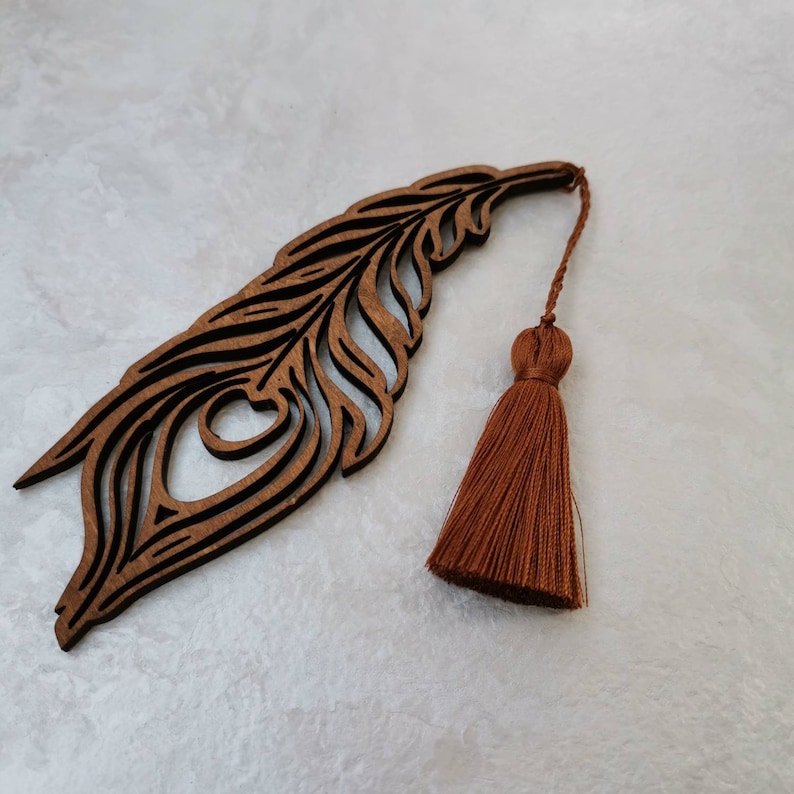 May include: A wooden bookmark shaped like a peacock feather with a brown tassel. The bookmark is intricately carved with details of the feather.
