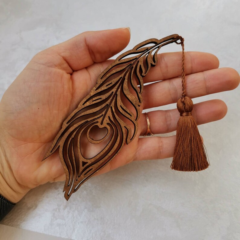 May include: A wooden peacock feather bookmark with a brown tassel. The bookmark is intricately carved with a detailed design of a peacock feather.