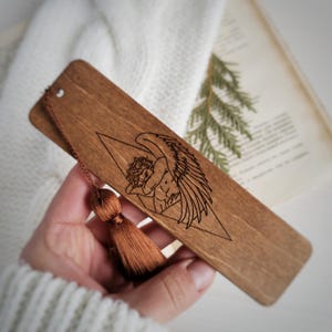 Peut inclure: Marque-page en bois avec une gravure détaillée d'un ange dans un triangle. Le marque-page est de couleur marron chaud et possède un pompon attaché. L'ange a de grandes ailes et est en position de repos. Le marque-page est tenu dans une main.