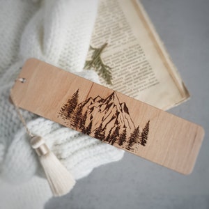 Könnte beinhalten: Ein Holz-Lesezeichen mit einem lasergravierten Design aus einer Bergkette und Bäumen. Das Lesezeichen hat einen Quasten dran.