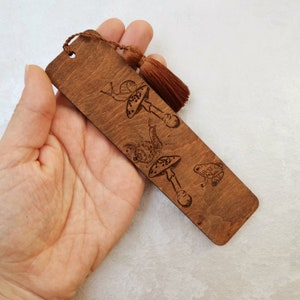 May include: A wooden bookmark with a brown tassel. The bookmark features a laser-engraved design of mushrooms, snails, and a frog.