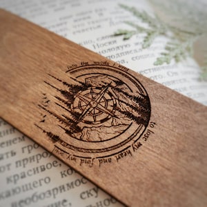 Può includere: Segnalibro in legno con un design a bussola e il testo "Into the woods I went to lose my roots".