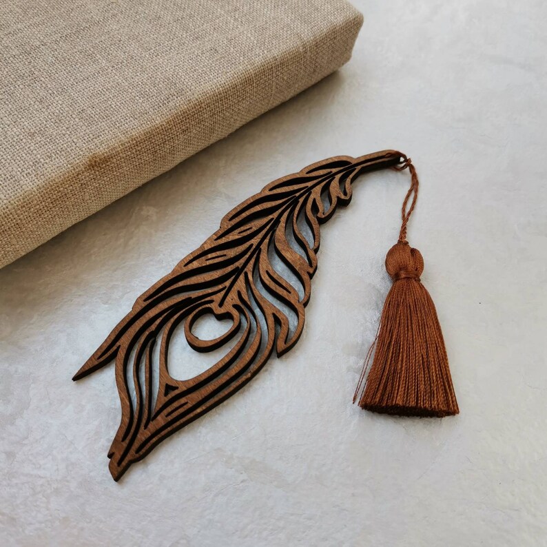 May include: A wooden peacock feather bookmark with a brown tassel. The bookmark is intricately carved with a detailed design.