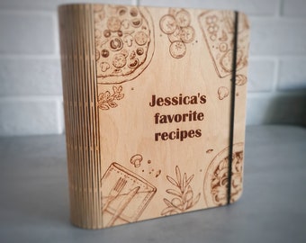 May include: A wooden recipe binder with a brown elastic band. The cover features a laser-engraved design of pizza, rosemary, and other food items. The text "Jessica's favorite recipes" is engraved on the cover.