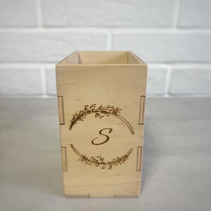 May include: A rectangular light-coloured wooden pencil holder featuring a laser-engraved design on the front. The design consists of a circular wreath made of thin branches and leaves, with a large, elegant capital letter S centred inside.