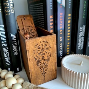 Könnte beinhalten: Holzkiste mit Scharnierdeckel, mit einem eingravierten Design einer Windrose, eines Baumes und Runen. Die Kiste ist geöffnet und zeigt einen Anhänger. Auf einem Regal mit Büchern und einer Kerze.