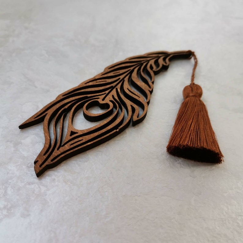 May include: A wooden peacock feather bookmark with a brown tassel. The bookmark is intricately carved and features a detailed eye design.