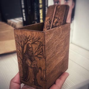 May include: A wooden box with a dark brown finish, featuring an engraved design of the Grim Reaper holding a book under a bare tree. The box is rectangular and holds several wooden cards. A spider is also engraved on the side.
