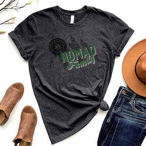 May include: A dark gray t-shirt with a green and black graphic design featuring a compass, mountains, and the text "Nomad Family".