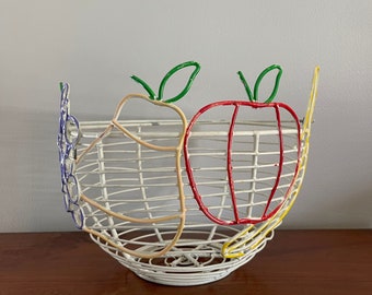 Può includere: Un cesto di frutta in filo bianco con un design di mela rossa, pera gialla e uva viola. Il cesto è realizzato in filo metallico e ha una finitura bianca.